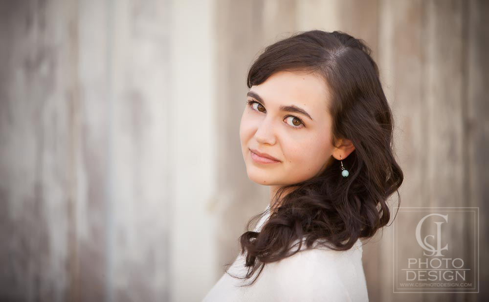 Senior girl in a white top and blue earrings against a rough clapboard background